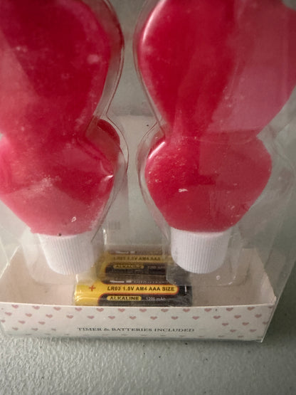 View of Included Batteries: Factory-sealed 2-pack box of At Home Red Heart LED Taper  Candles, showcasing two stacked heart-shaped flameless sticks with included batteries.