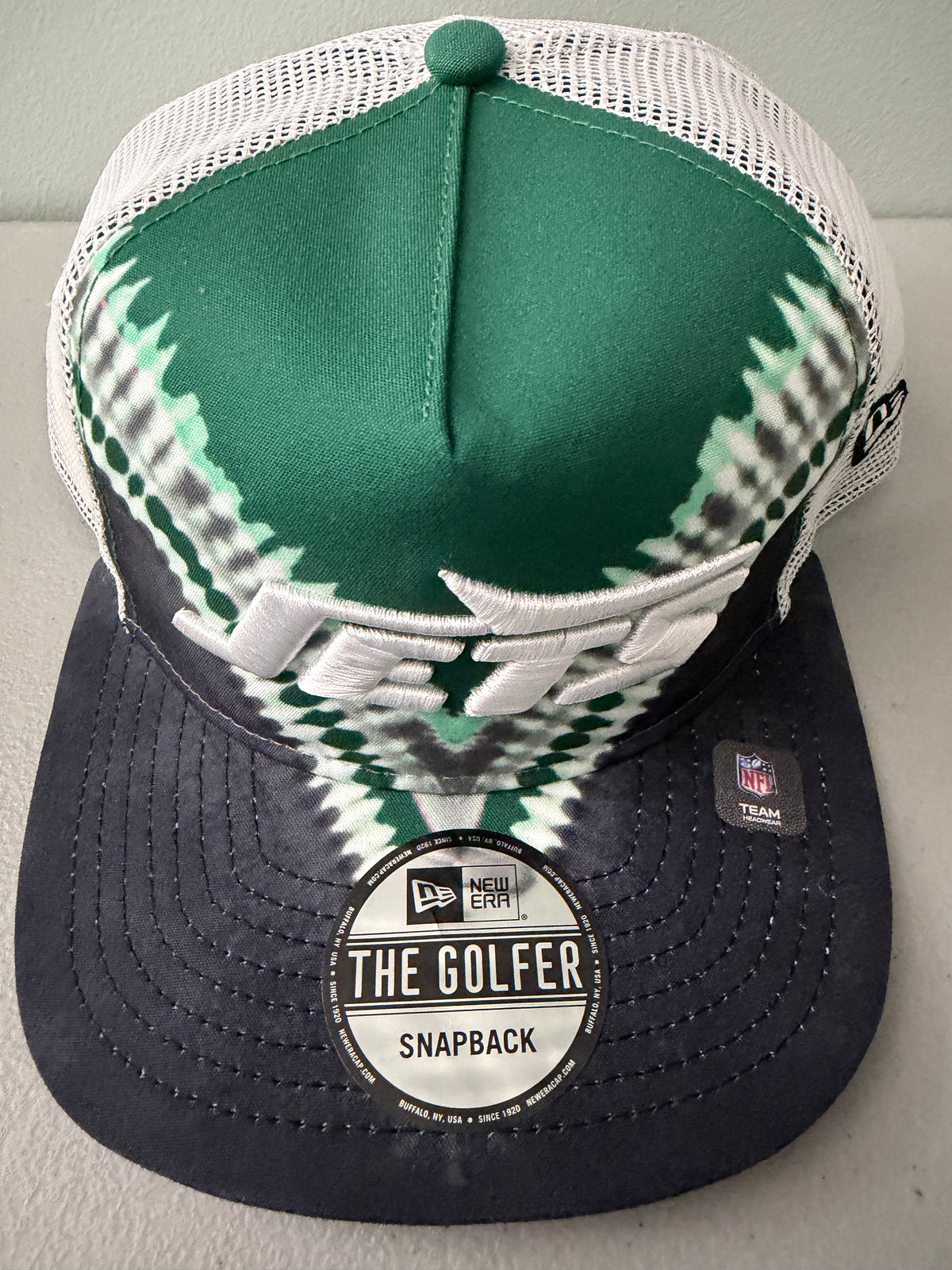 Official Brand Sticker: Front profile shot of a brand new New Era New York Jets The Golfer snapback hat featuring a green and white sublimated tie dye front panel, premium embroidered white jets logo, gray structured brim, and breathable white mesh trucker backing. 