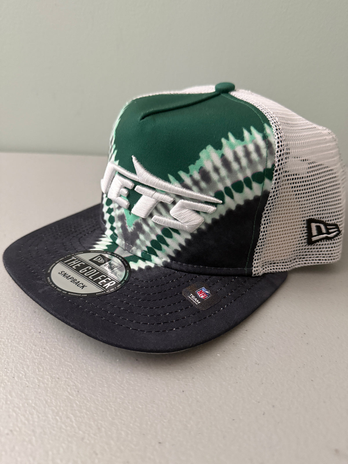 Angle View: Front profile shot of a brand new New Era New York Jets The Golfer snapback hat featuring a green and white sublimated tie dye front panel, premium embroidered white jets logo, gray structured brim, and breathable white mesh trucker backing. 