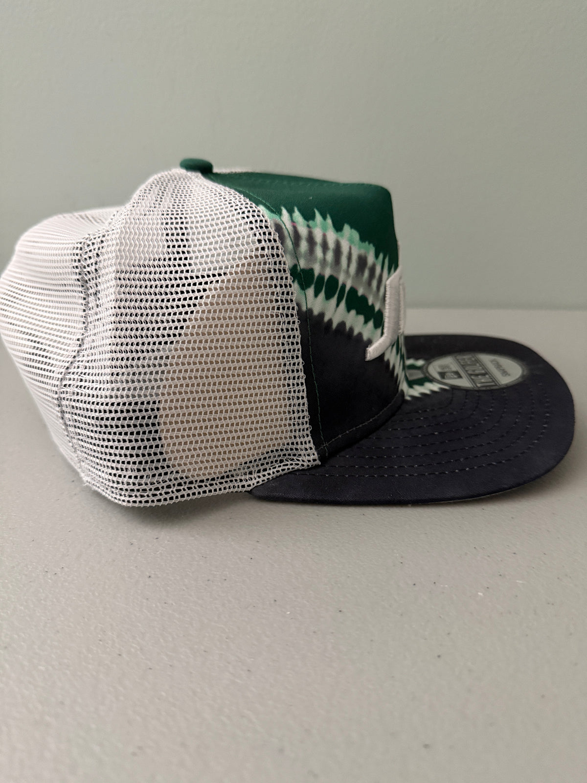 Other Side View: Front profile shot of a brand new New Era New York Jets The Golfer snapback hat featuring a green and white sublimated tie dye front panel, premium embroidered white jets logo, gray structured brim, and breathable white mesh trucker backing. 