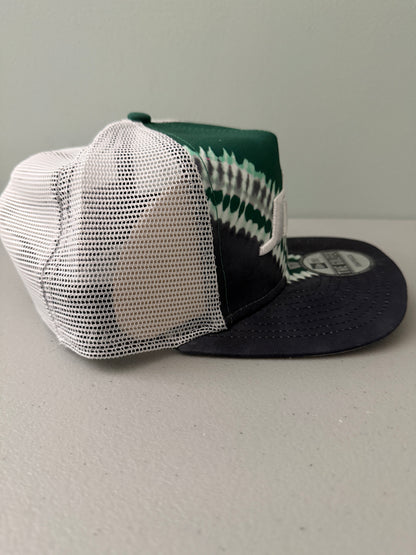 Other Side View: Front profile shot of a brand new New Era New York Jets The Golfer snapback hat featuring a green and white sublimated tie dye front panel, premium embroidered white jets logo, gray structured brim, and breathable white mesh trucker backing. 
