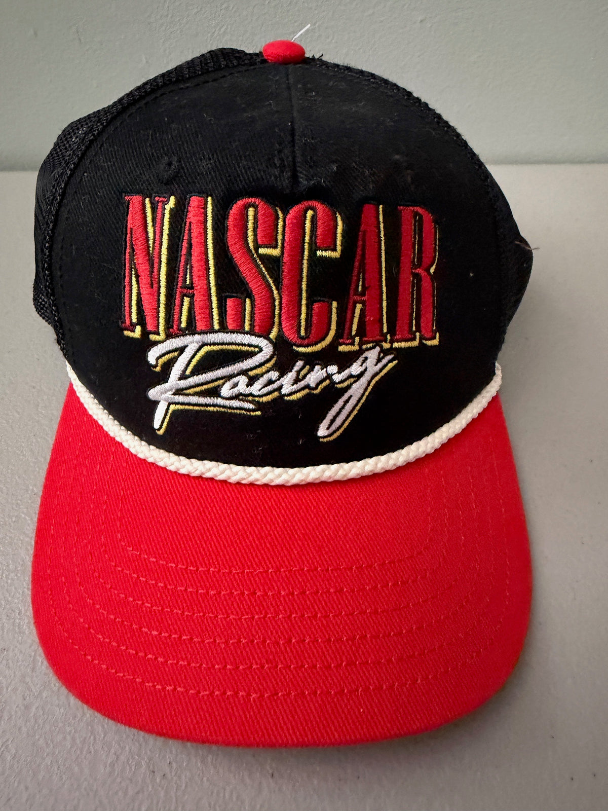 Brand new 2026 NASCAR racing rope trucker hat in black and red, featuring a vibrant embroidered front logo, white braided rope trim, and adjustable snapback closure