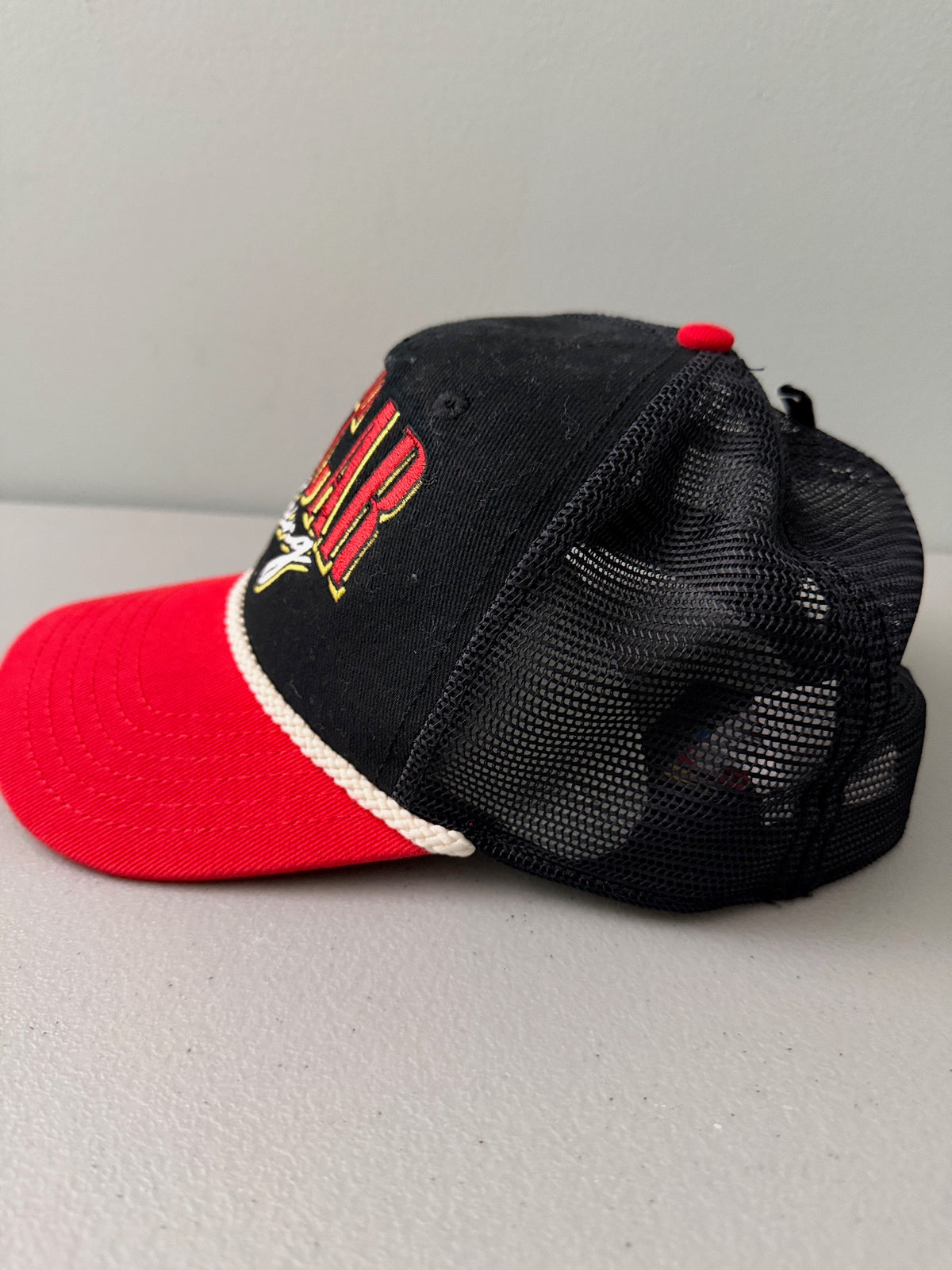 Side View: Brand new 2026 NASCAR racing rope trucker hat in black and red, featuring a vibrant embroidered front logo, white braided rope trim, and adjustable snapback closure