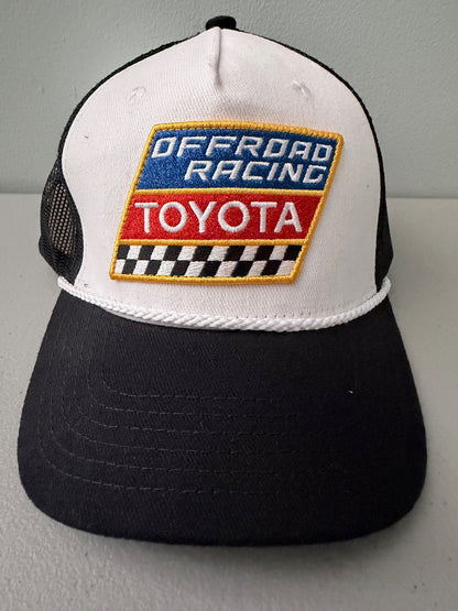 Brand new Toyota Off-Road Racing rope trucker hat in black and white, featuring a large embroidered retro shield logo patch, checkerboard racing stripes, and a braided white rope detail on a snapback cap from Bell's Bargain Barn.