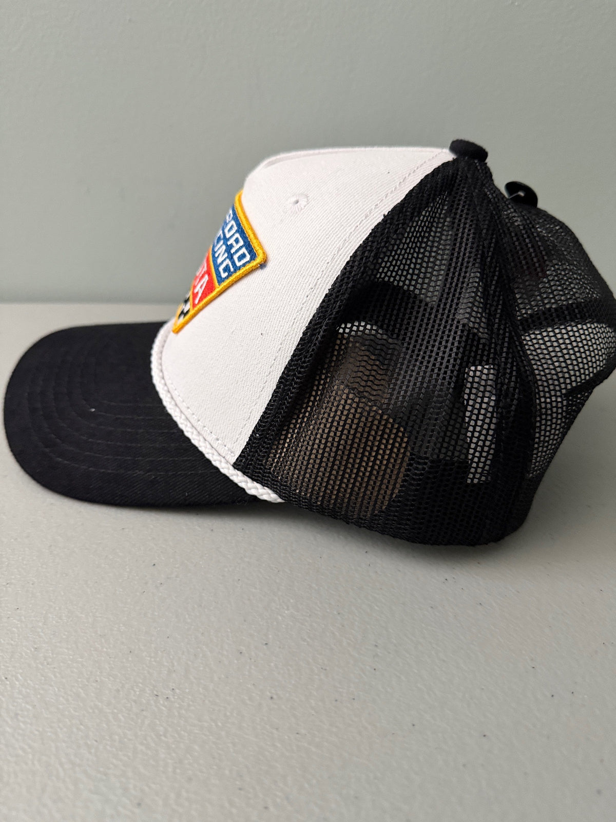 Side View: Brand new Toyota Off-Road Racing rope trucker hat in black and white, featuring a large embroidered retro shield logo patch, checkerboard racing stripes, and a braided white rope detail on a snapback cap from Bell's Bargain Barn.