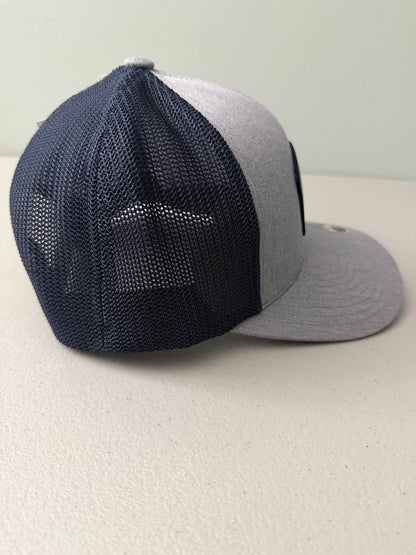 Right Side View: Brand new Travis Mathew Bush Wood CC 1980 Caddy Day Golf Tournament Champion trucker hat featuring a heather gray structured crown, an embroidered champion patch, dark blue mesh backing, and a gold FlexFit 110 sticker from Bell's Bargain Barn.