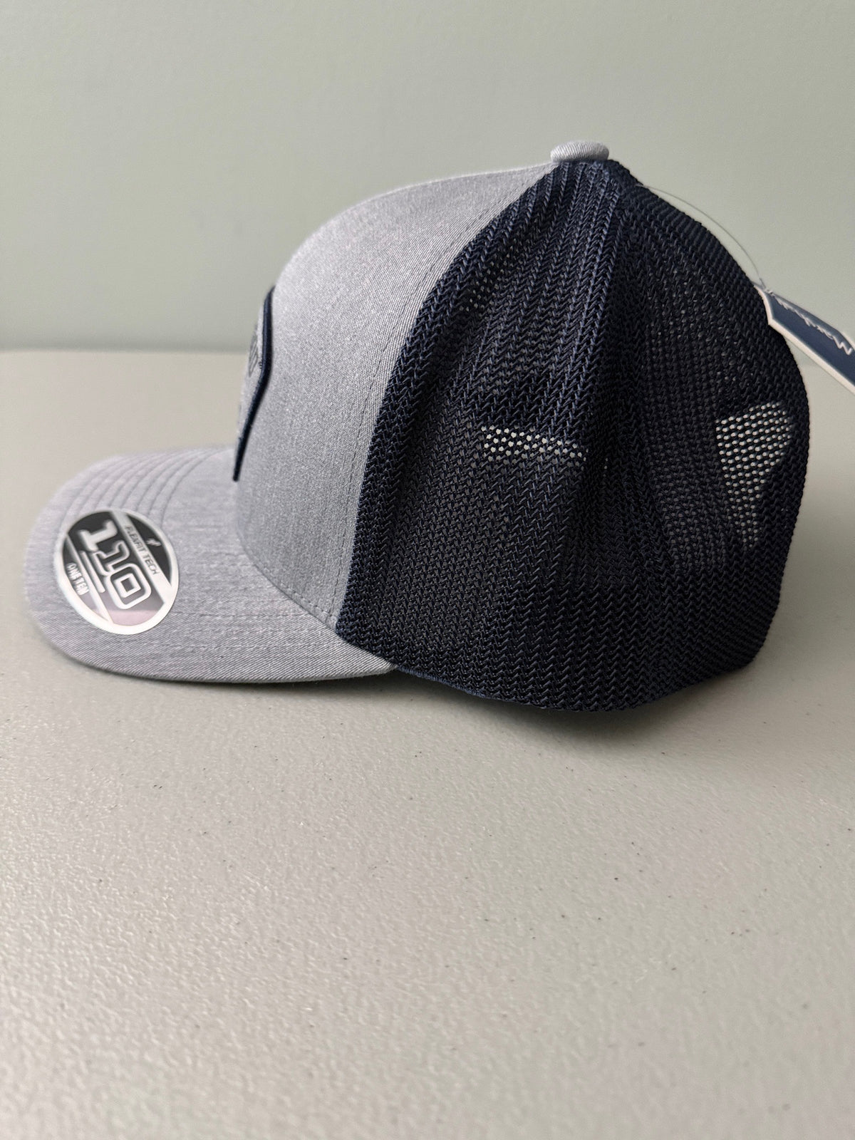 Left Side View: Brand new Travis Mathew Bush Wood CC 1980 Caddy Day Golf Tournament Champion trucker hat featuring a heather gray structured crown, an embroidered champion patch, dark blue mesh backing, and a gold FlexFit 110 sticker from Bell's Bargain Barn.