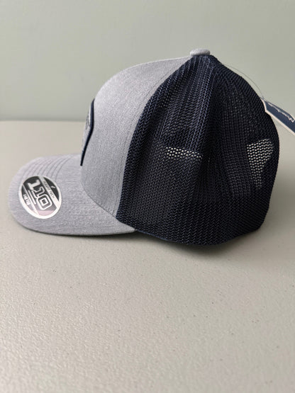 Left Side View: Brand new Travis Mathew Bush Wood CC 1980 Caddy Day Golf Tournament Champion trucker hat featuring a heather gray structured crown, an embroidered champion patch, dark blue mesh backing, and a gold FlexFit 110 sticker from Bell's Bargain Barn.