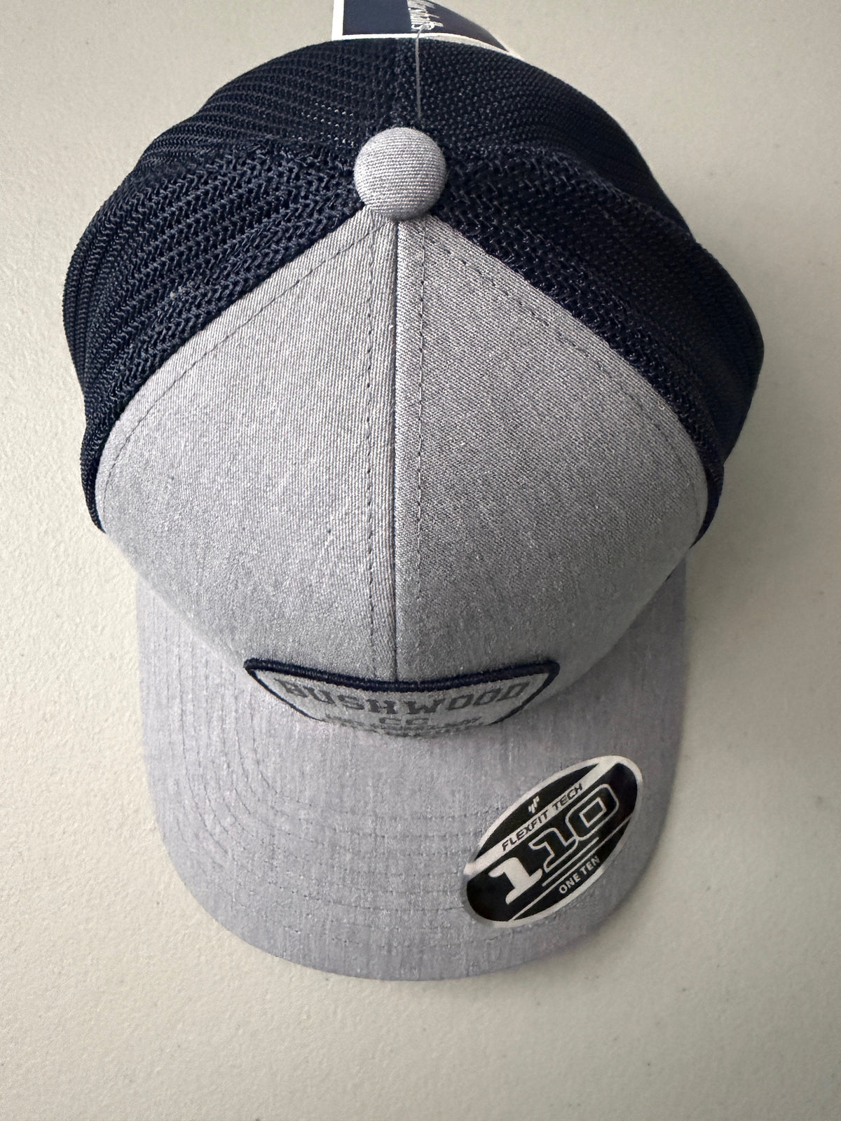 Top View: Brand new Travis Mathew Bush Wood CC 1980 Caddy Day Golf Tournament Champion trucker hat featuring a heather gray structured crown, an embroidered champion patch, dark blue mesh backing, and a gold FlexFit 110 sticker from Bell's Bargain Barn.