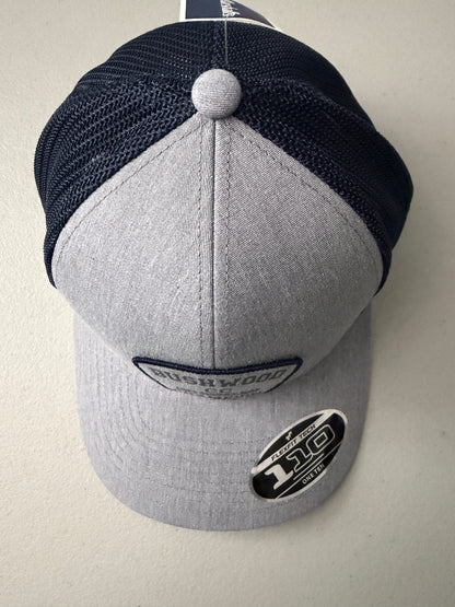 Top View: Brand new Travis Mathew Bush Wood CC 1980 Caddy Day Golf Tournament Champion trucker hat featuring a heather gray structured crown, an embroidered champion patch, dark blue mesh backing, and a gold FlexFit 110 sticker from Bell's Bargain Barn.