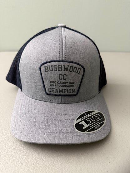 Brand new Travis Mathew Bush Wood CC 1980 Caddy Day Golf Tournament Champion trucker hat featuring a heather gray structured crown, an embroidered champion patch, dark blue mesh backing, and a gold FlexFit 110 sticker from Bell's Bargain Barn.