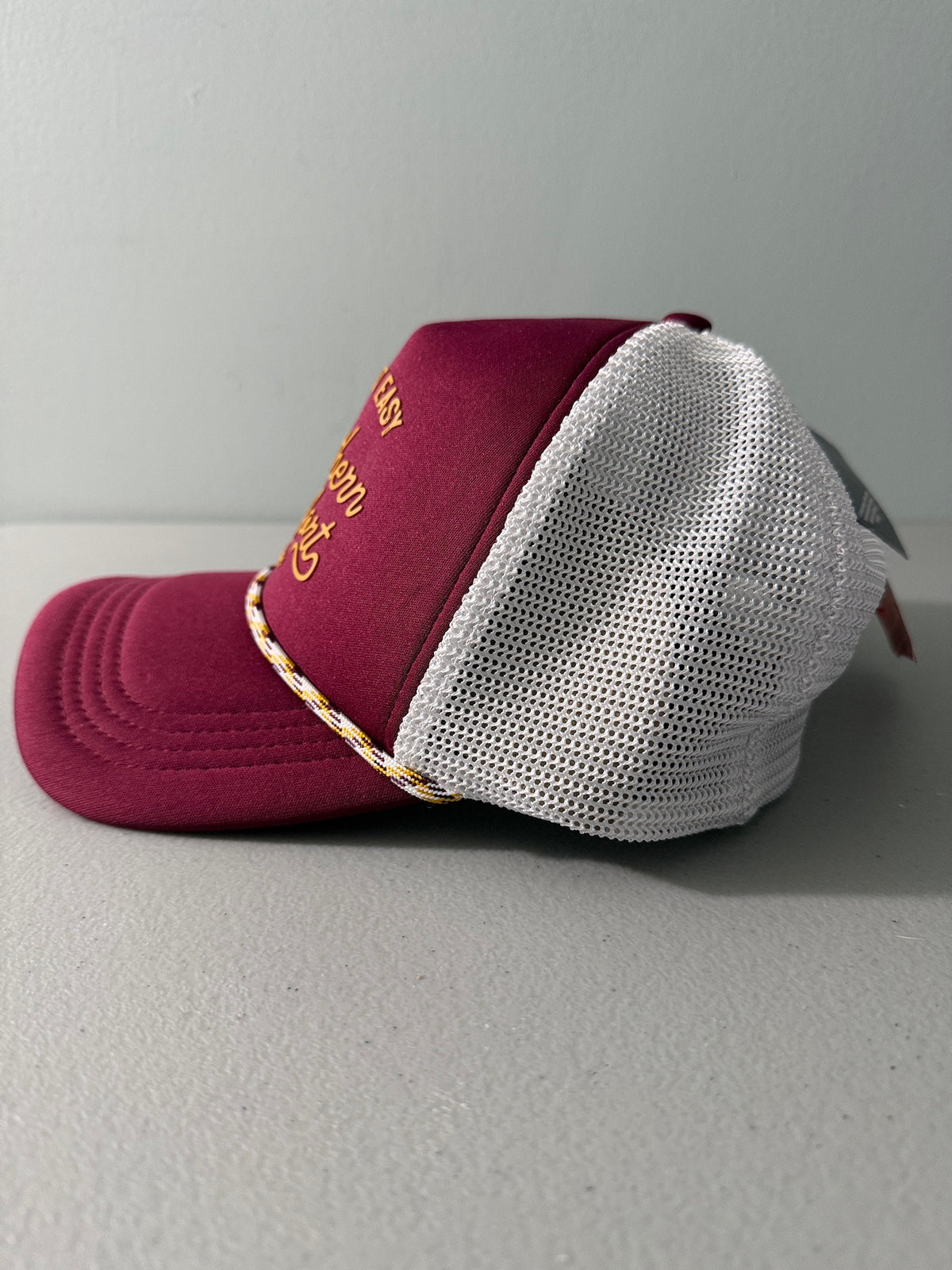 Side View: Brand new Southern Comfort Take It Easy mesh trucker hat featuring a structured maroon front foam panel, raised gold cursive lettering, white mesh back panels, a patterned front visor accent rope, and an adjustable snapback closre from Bell's Bargain Barn. 