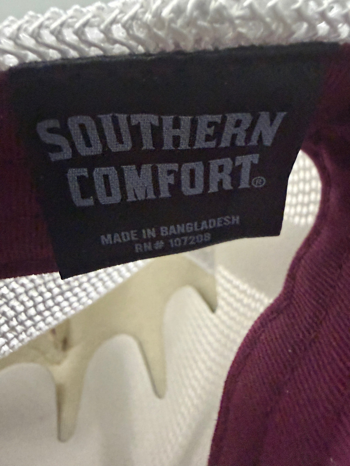 Product Brand Tag: Brand new Southern Comfort Take It Easy mesh trucker hat featuring a structured maroon front foam panel, raised gold cursive lettering, white mesh back panels, a patterned front visor accent rope, and an adjustable snapback closre from Bell's Bargain Barn. 