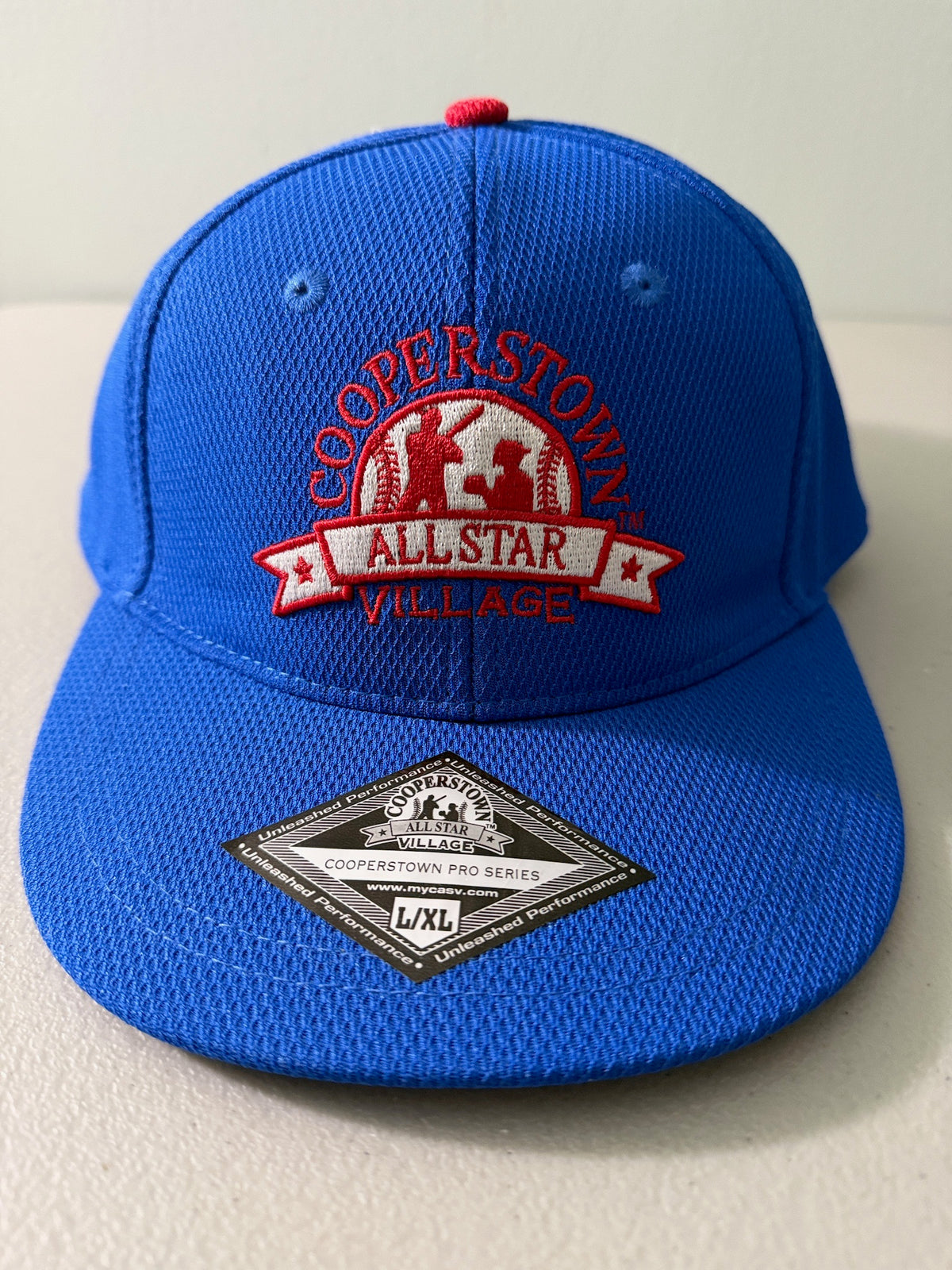 Brand new royal blue Cooperstown All Star Pro Village Pro Series stretch fit baseball hat featuring an embroidered red and white tournament patch, a diamond-shaped L/XL silver visor sticker, and an athletic mesh-textured crown from Bell's Bargain Barn. 