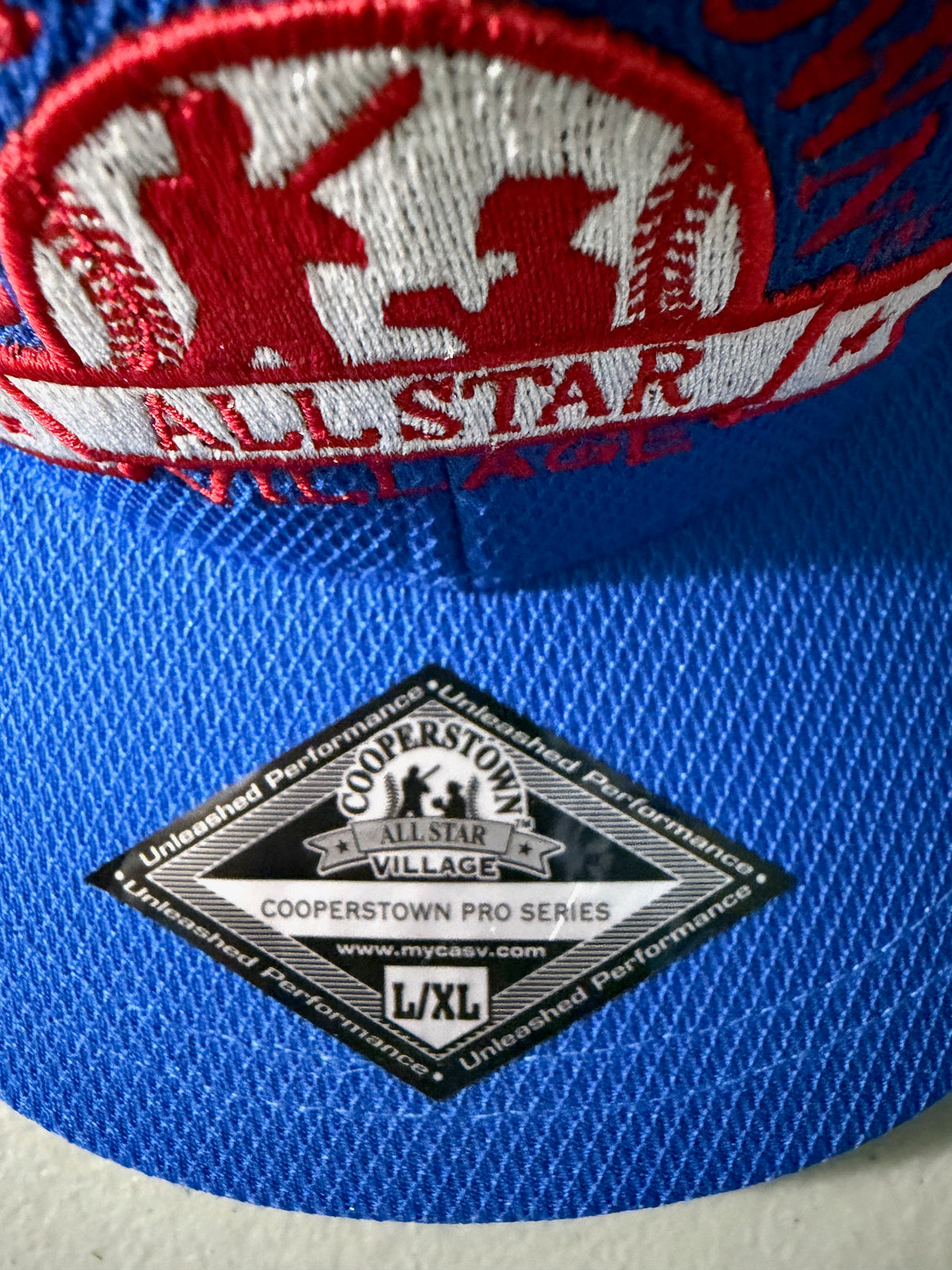 Silver Visor Sticker View: Brand new royal blue Cooperstown All Star Pro Village Pro Series stretch fit baseball hat featuring an embroidered red and white tournament patch, a diamond-shaped L/XL silver visor sticker, and an athletic mesh-textured crown from Bell's Bargain Barn. 
