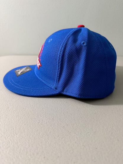 Side View: Brand new royal blue Cooperstown All Star Pro Village Pro Series stretch fit baseball hat featuring an embroidered red and white tournament patch, a diamond-shaped L/XL silver visor sticker, and an athletic mesh-textured crown from Bell's Bargain Barn. 