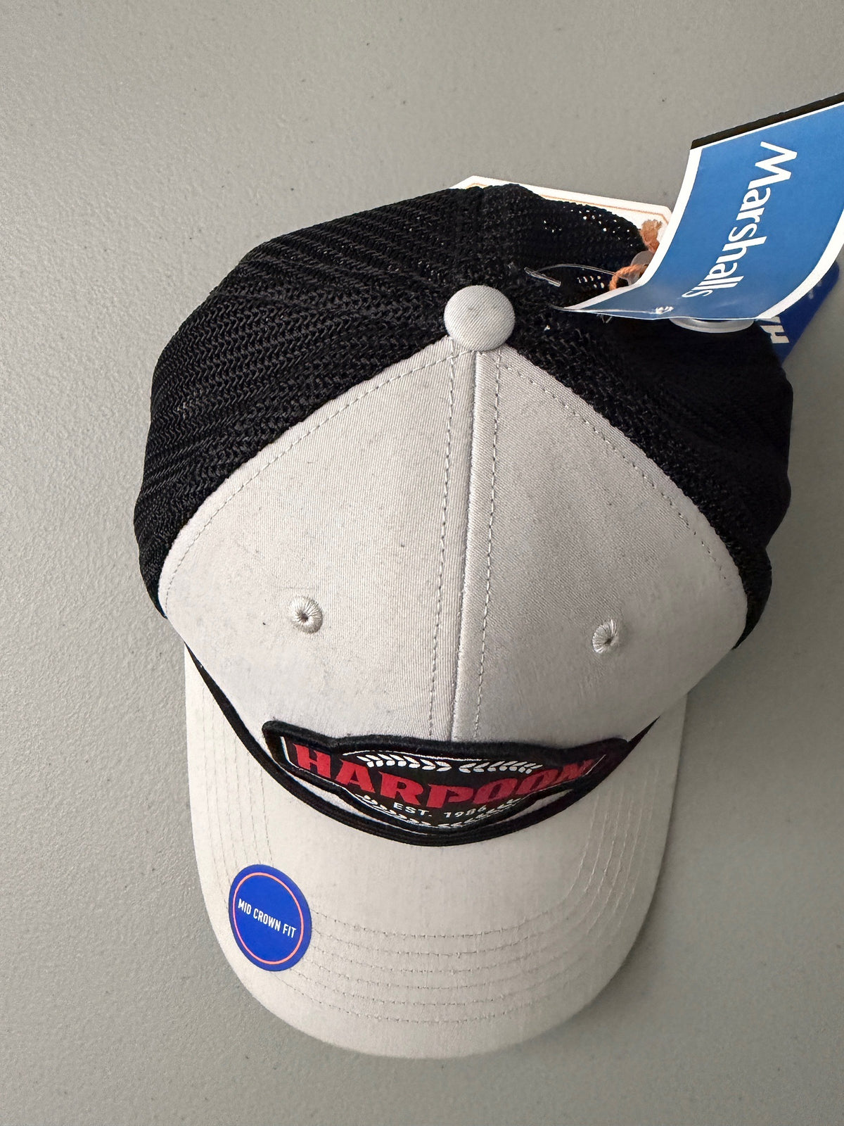 Top View: Brand new Harpoon Brewery Est. 198 mid crown fit trucker hat featuring a structured off-white front panel with a black embroidered logo patch, black mesh backing, a thin black visor rope, and a blue brim sticker from bell's Bargain Barn.