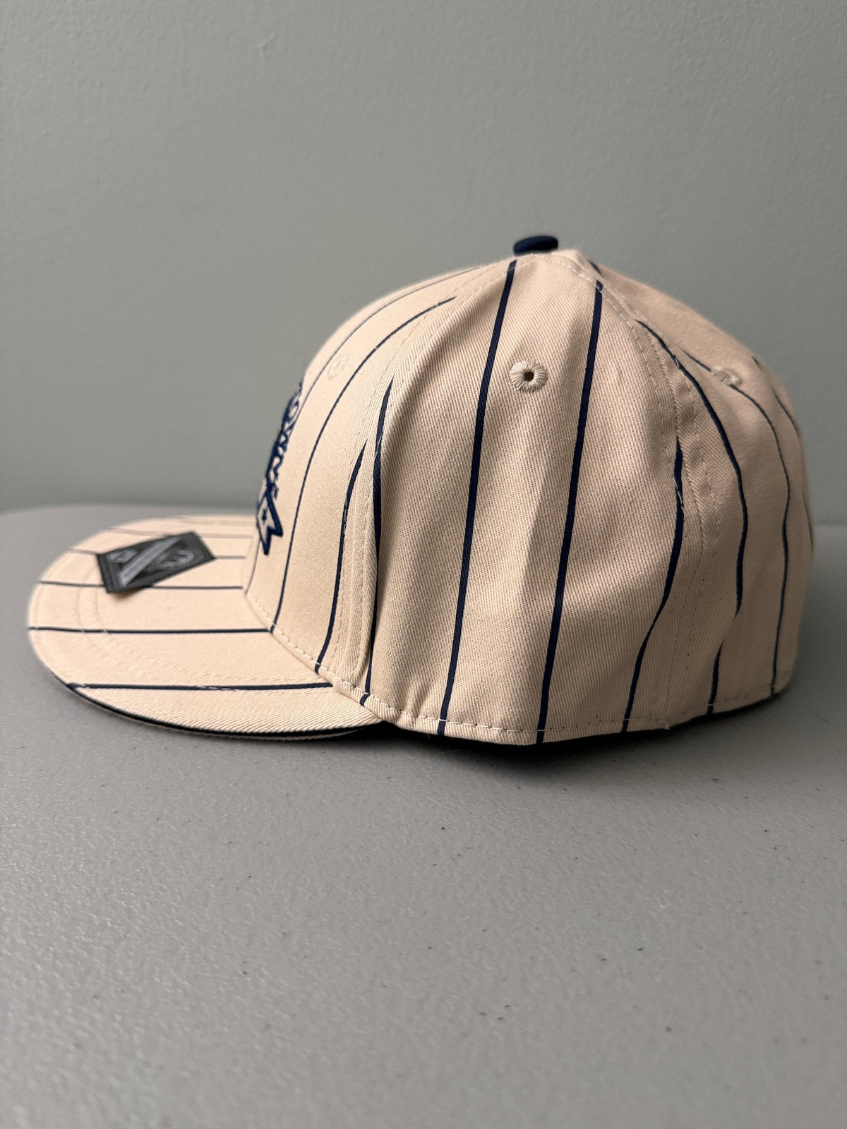 Side View: Brand new cream Cooperstown All Star Village Pro Series stretch fit baseball hat with navy blue pinstripes, featuring an embroidered navy tournament logo patch and a silver diamond-shaped L/XL visor sticker from Bell's Bargain Barn.  