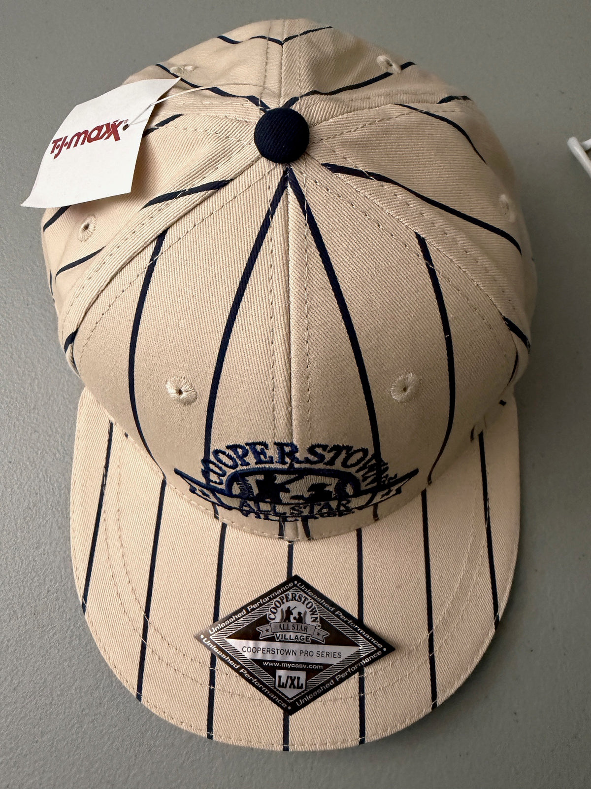 Top View: Brand new cream Cooperstown All Star Village Pro Series stretch fit baseball hat with navy blue pinstripes, featuring an embroidered navy tournament logo patch and a silver diamond-shaped L/XL visor sticker from Bell's Bargain Barn.  