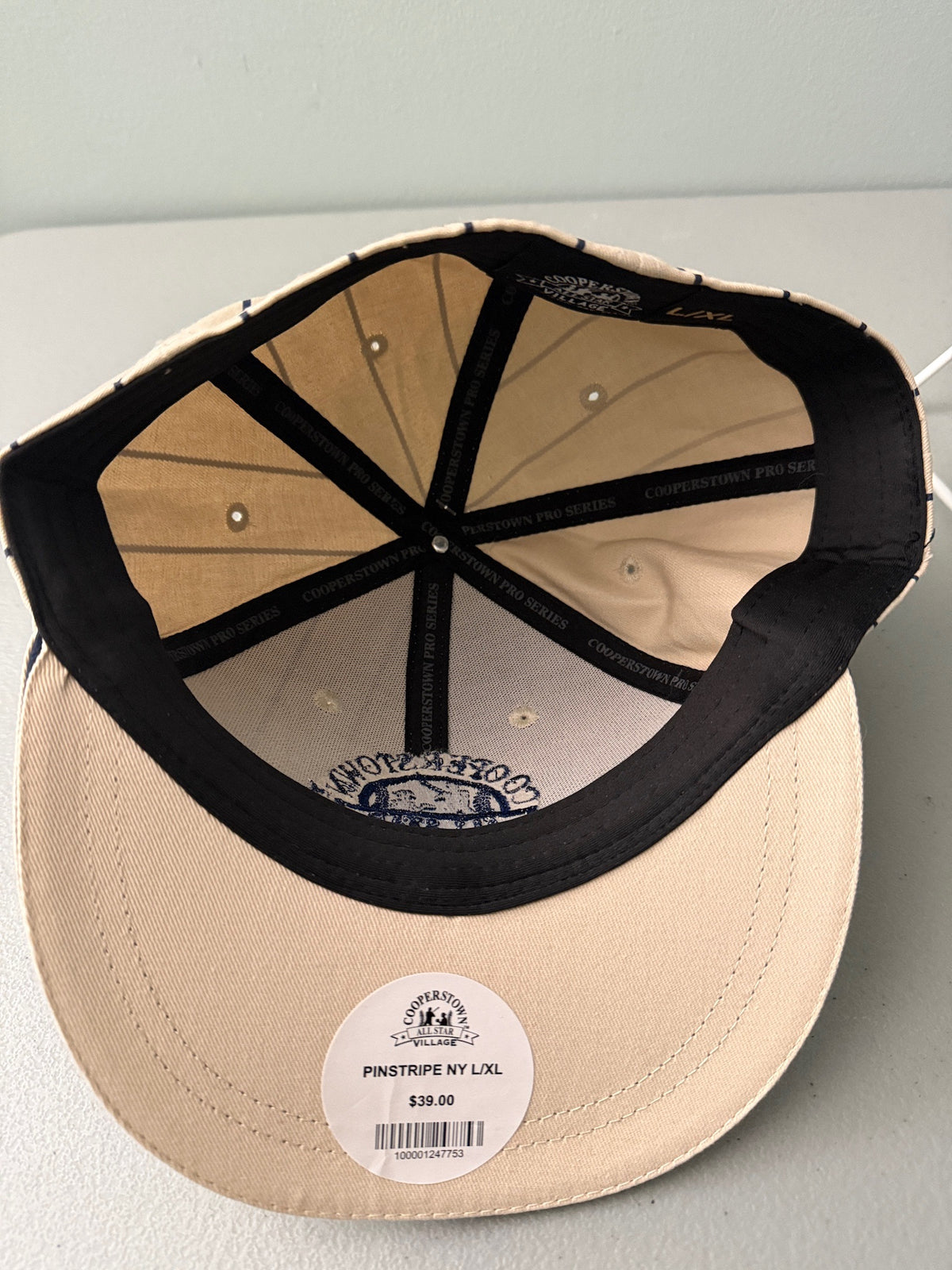 Inside View: Brand new cream Cooperstown All Star Village Pro Series stretch fit baseball hat with navy blue pinstripes, featuring an embroidered navy tournament logo patch and a silver diamond-shaped L/XL visor sticker from Bell's Bargain Barn.  