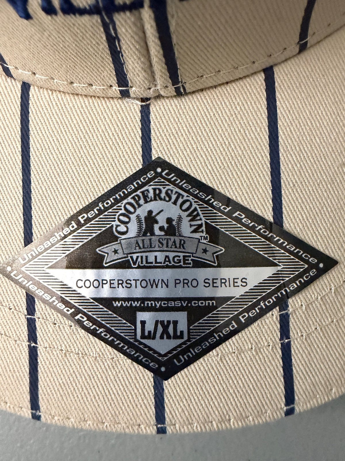 Silver Diamond Shaped L/XL Visor Sticker: Brand new cream Cooperstown All Star Village Pro Series stretch fit baseball hat with navy blue pinstripes, featuring an embroidered navy tournament logo patch and a silver diamond-shaped L/XL visor sticker from Bell's Bargain Barn.  