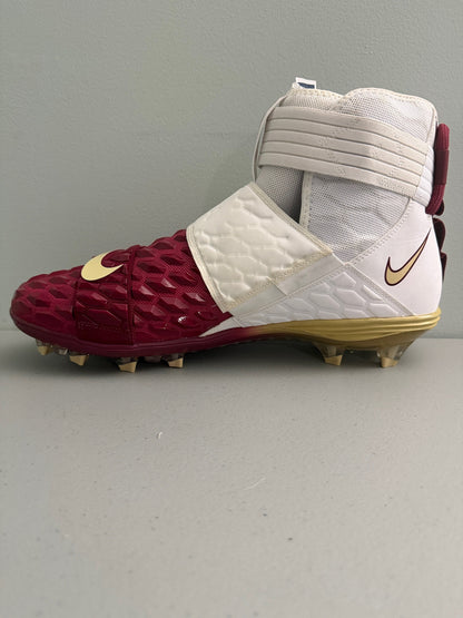A Nike high-top football cleat in maroon, white, and gold, featuring a textured maroon toe box, white mesh and quilted panels, and gold Nike Swooshes.