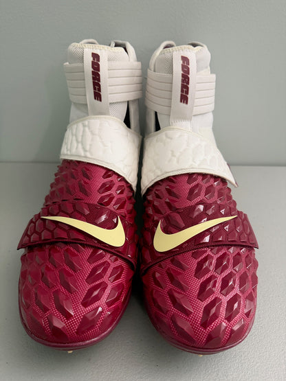 A pair of Nike Force 10 high-top Football cleats in a deep maroon red and white colorway, featuring a textured honeycomb pattern on the toe box and a yellow Nike Swoosh on the strap. 