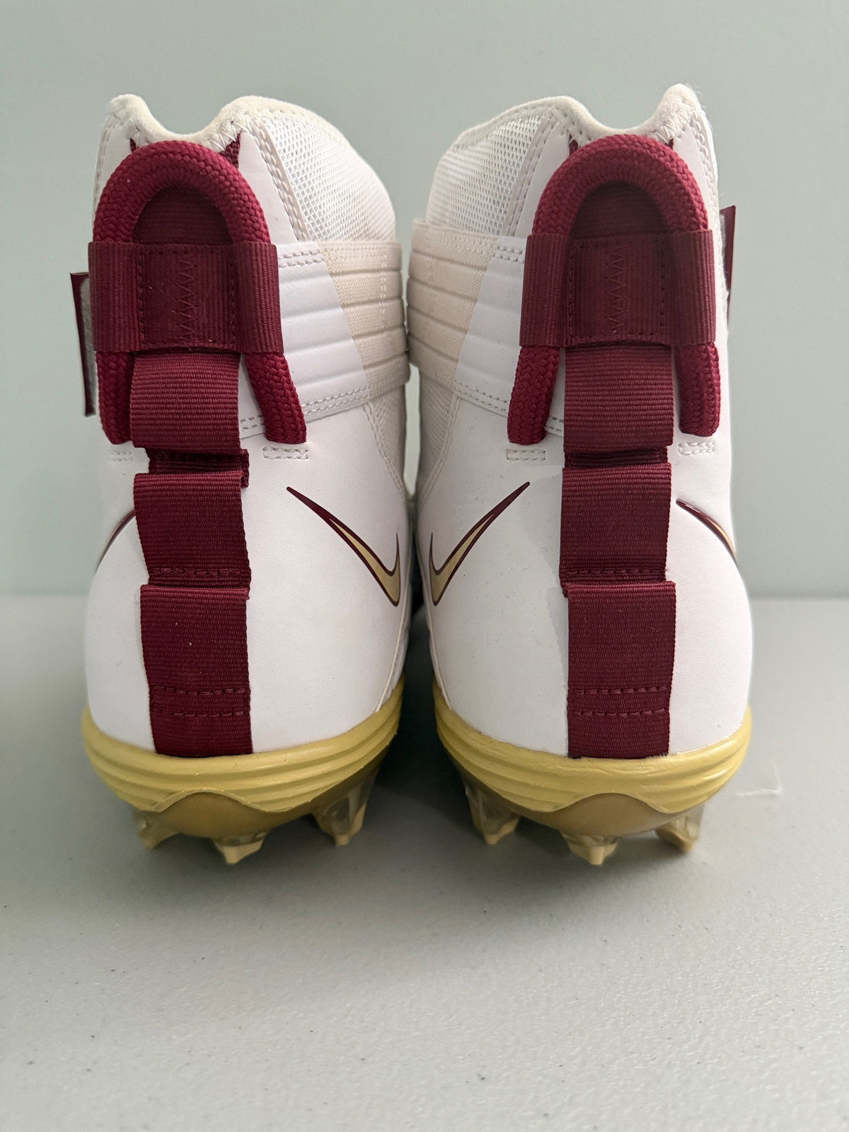 A pair of Nike high-top football cleats shown from the back, featuring a white leather and mesh upper with maroon heel straps, a maroon Nike swoosh, and gold cleats in rare size US 18 (Big & Tall).