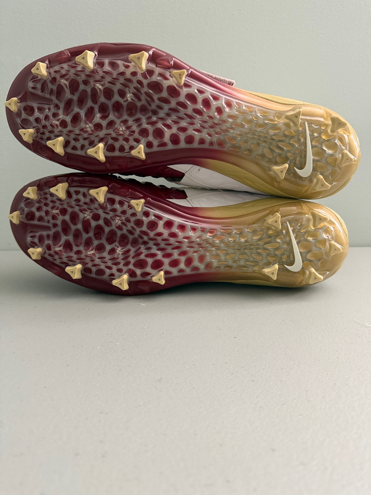 A pair of Nike turf Cleats shown from the sole side, featuring a translucent maroon and gold gradient with gold studs and a white Nike Swoosh. Rare big & tall size 18. 