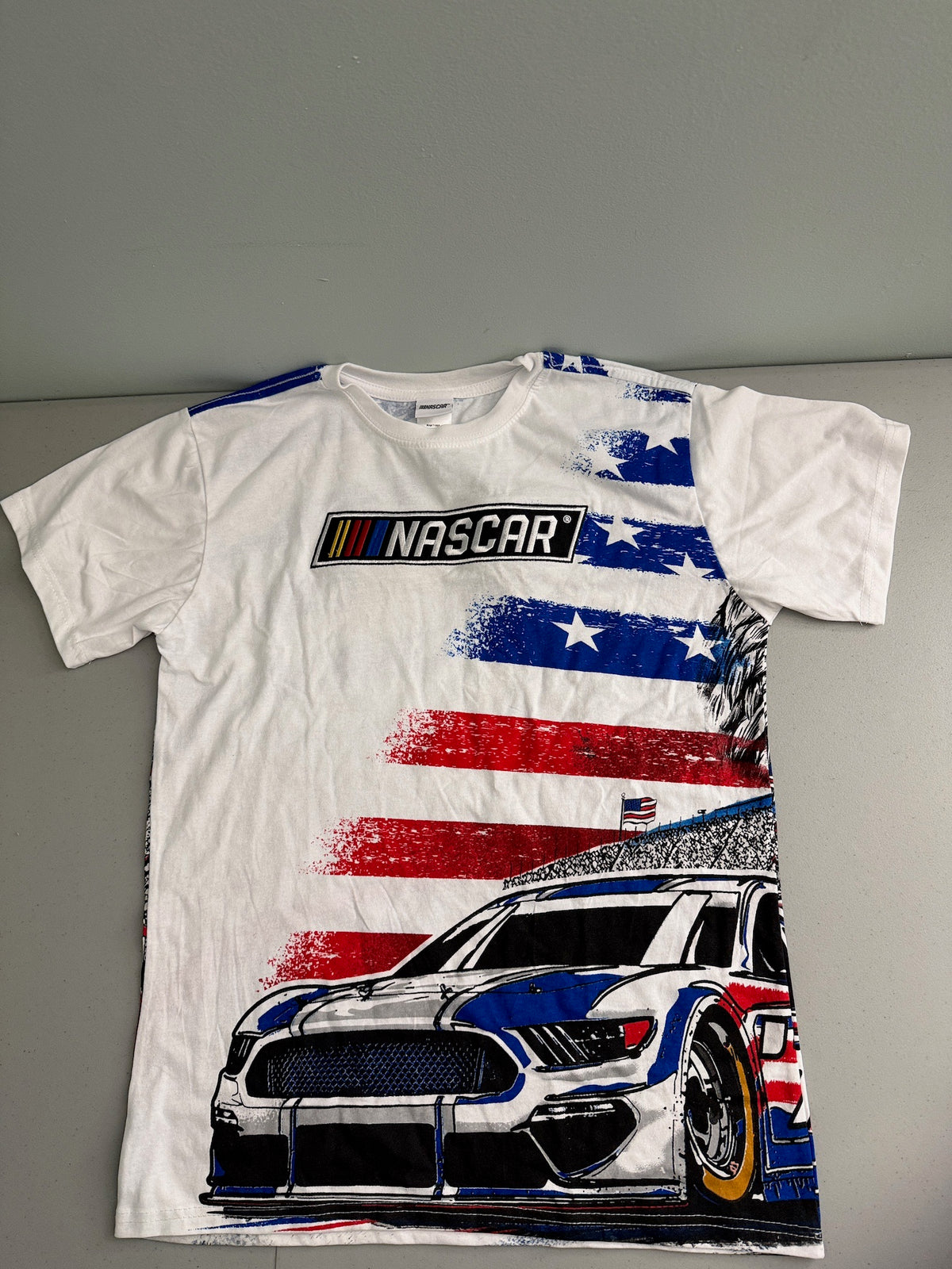 Front View: Officially licensed white NASCAR men's graphic t-shirt laid flat featuring an American flag design and blue race car print on the chest.
