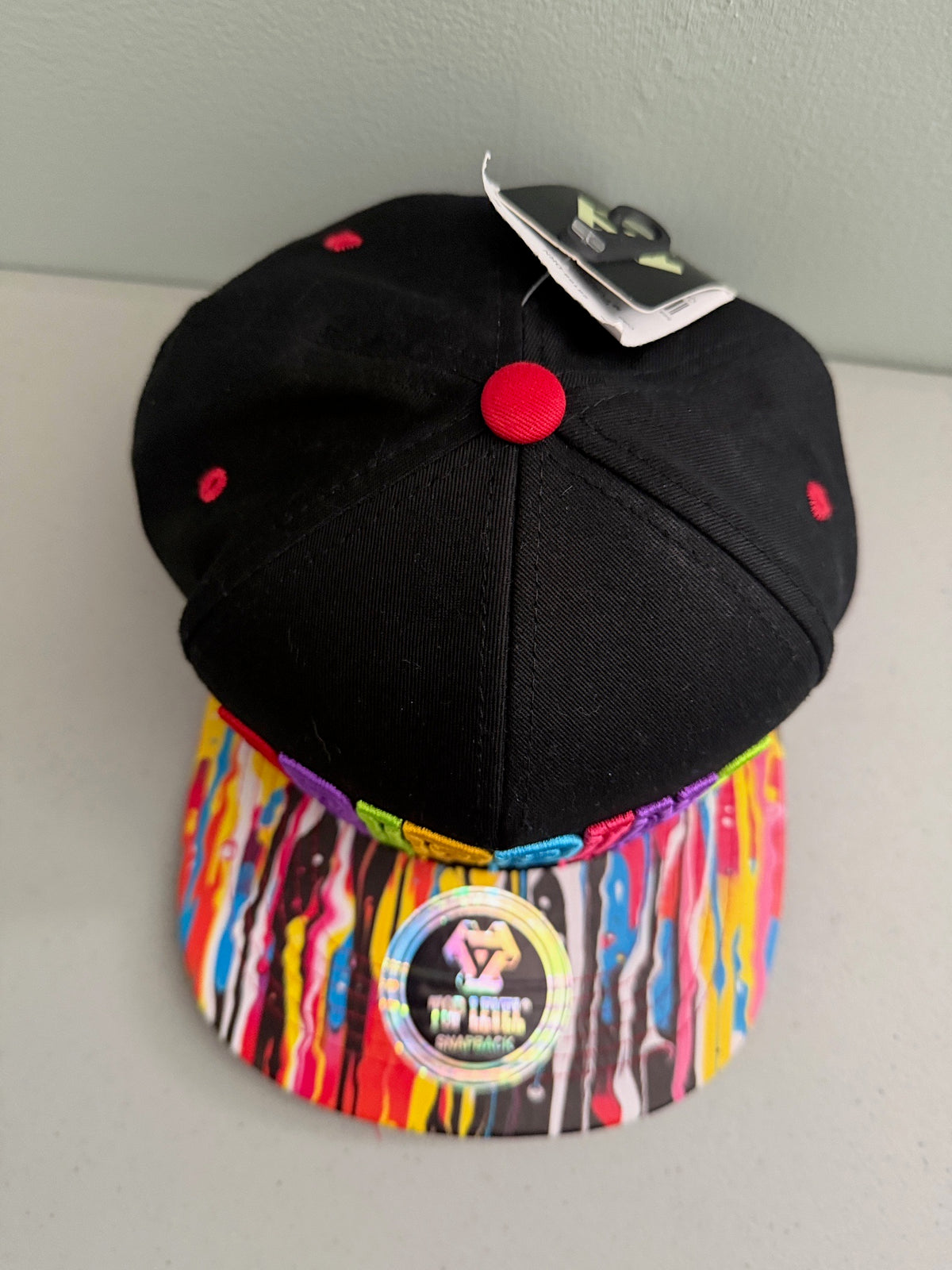 Top-down view of the black Top Level DRIPPING streetwear cap, highlighting the six-panel crown construction, top button, and the full layout of the vibrant multicolor paint splatter design across the flat bill.